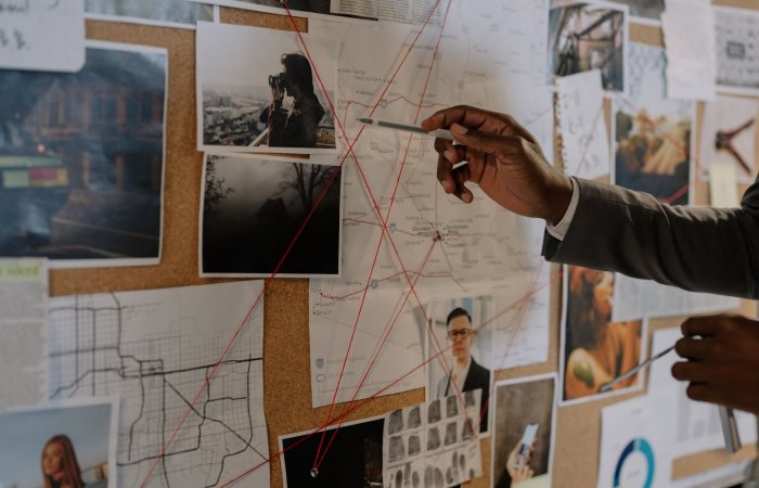 Detective examines a corkboard with maps and photos to solve a mystery.